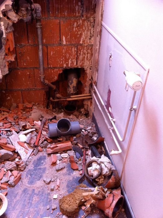 Scène de démolition avec mur en briques apparentes, tuyaux, débris au sol. Mur blanc avec radiateur.