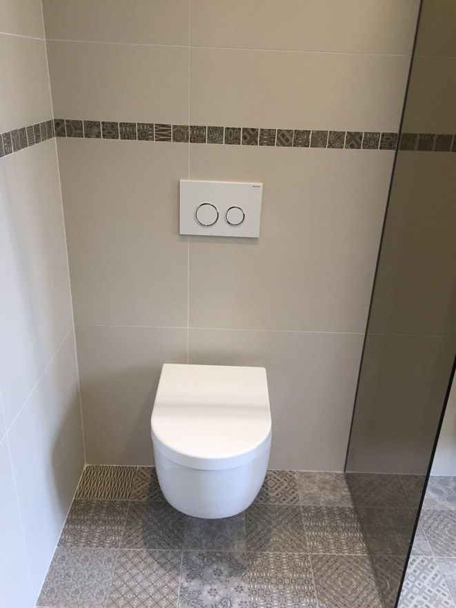 Toilettes murales blanches dans une petite salle de bain avec des murs en carrelage clair, un sol à motifs et une plaque de chasse d'eau à deux boutons.
