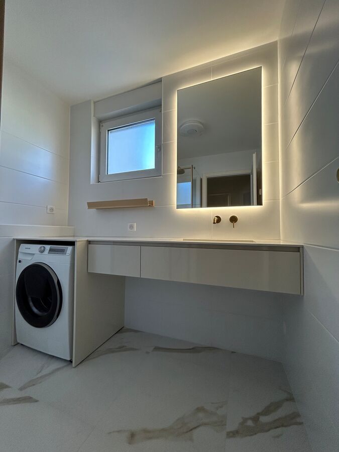 Une salle de bain avec machine à laver, lavabo, miroir et fenêtre.
