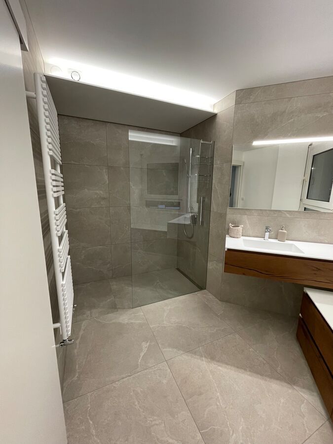 Salle de bain moderne avec douche en verre, carrelage gris clair, vanité en bois et radiateur blanc.