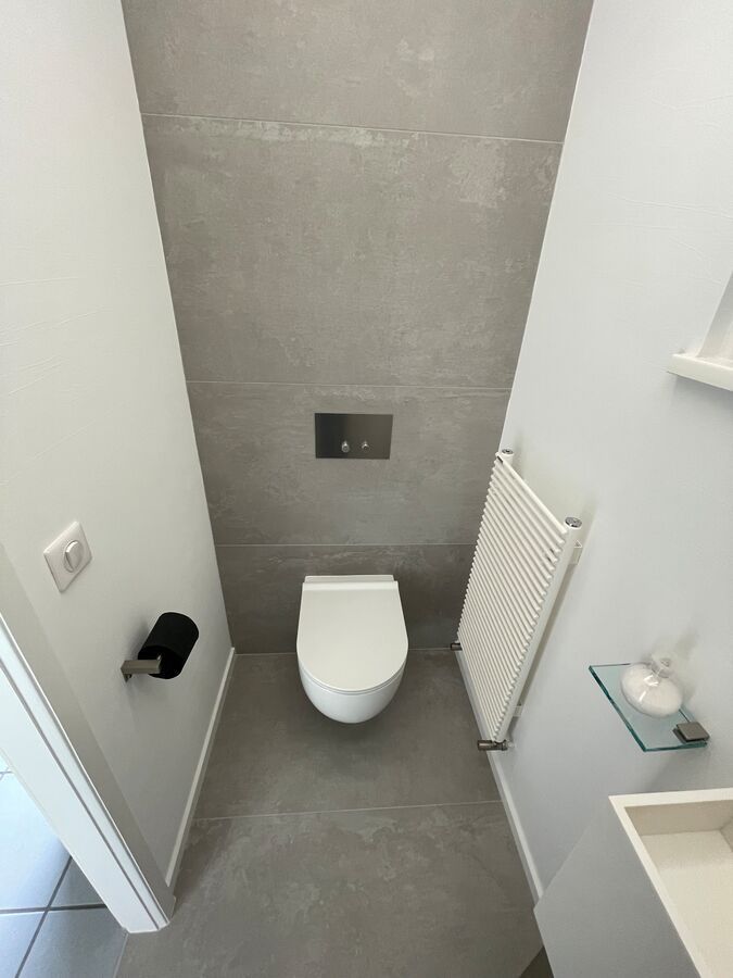 Salle de bain moderne et étroite avec carrelage gris au mur derrière les toilettes. Lavabo et radiateur blancs inclus.