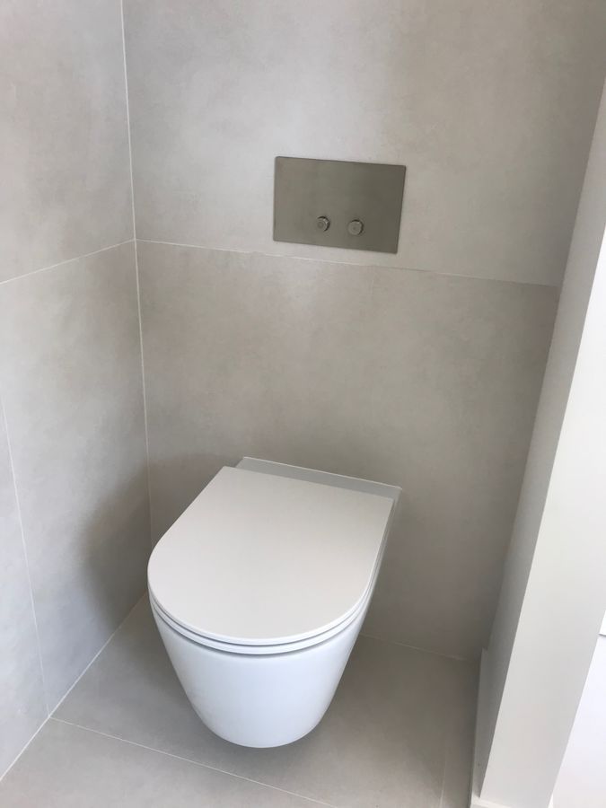 Toilettes murales modernes dans une salle de bain aux tons neutres. Dotées d'un siège blanc et de boutons de chasse d'eau muraux.