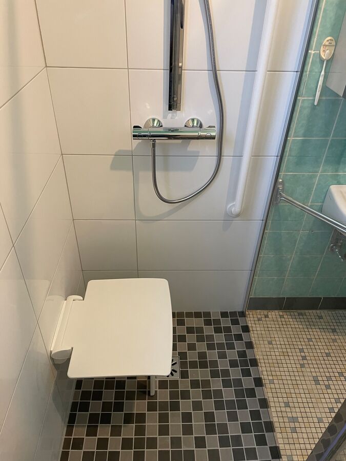 Cabine de douche avec siège rabattable, barre d'appui et douchette à main, carrelage blanc et carrelage au sol noir/gris.