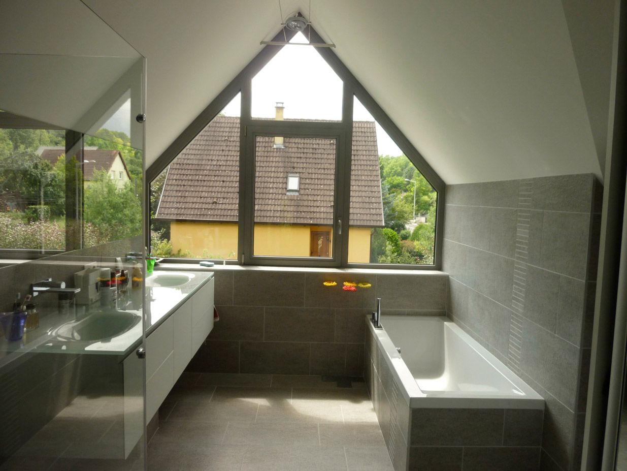 Salle de bain moderne avec plafond en pente, grande fenêtre triangulaire avec vue sur une maison et la nature.
