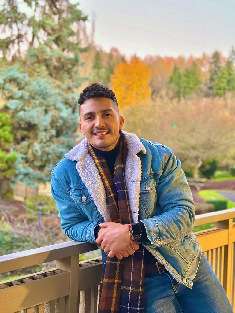 Man leaning on railing, wearing denim jacket with faux fur, scarf. Outdoors with fall foliage.