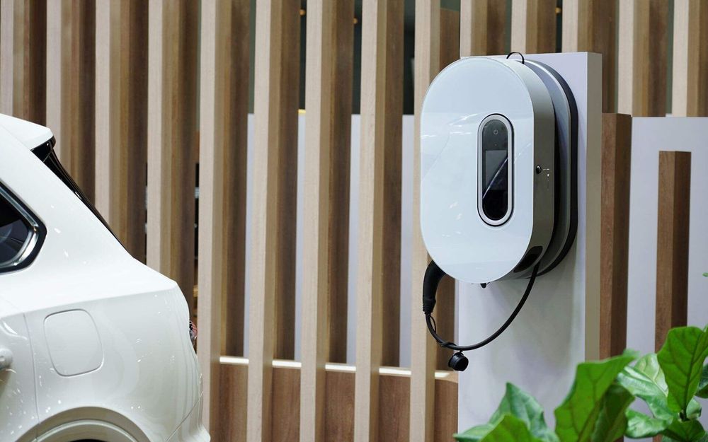 Electric car charging station beside a white car, with a wooden fence and plants in the background.