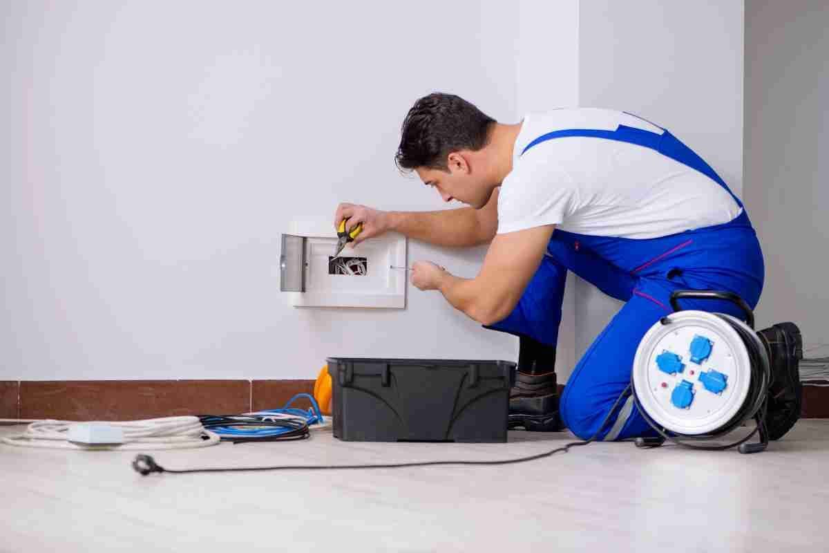 Electrician kneeling by a wall panel, wiring a white outlet box beside tools and cable reels.