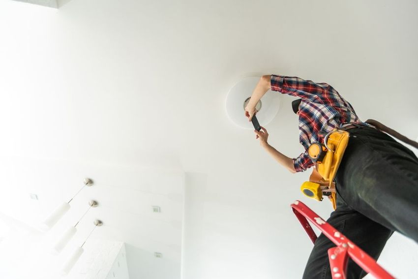 Worker on a red ladder installing a ceiling light in a white room