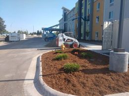 Construction equipment, including a boom lift and a skid steer, parked near a multi-story building with landscaping work.