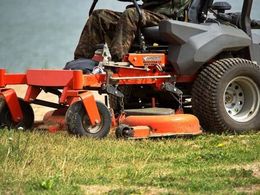 A person in camouflage pants operates an orange zero-turn lawnmower across a grassy field near water.