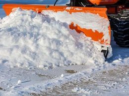 An orange snowplow blade attached to a vehicle pushes a large mound of snow across a snowy surface.