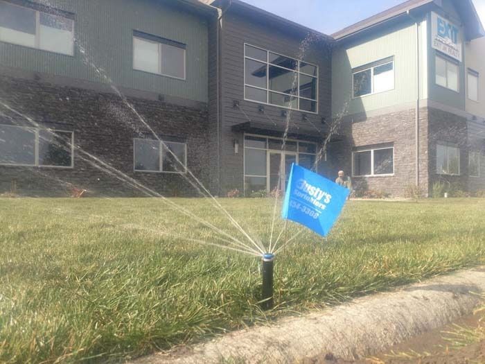 A lawn sprinkler sprays water in front of a commercial building with a blue advertising flag stuck in the grass.