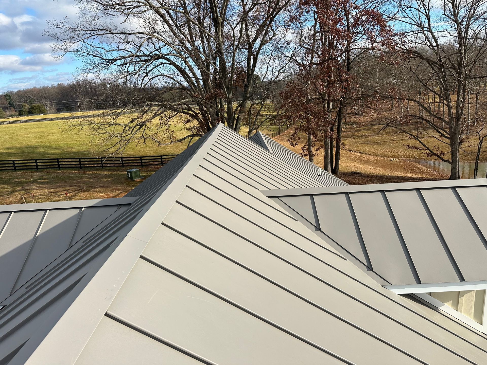 Gray metal roof on a house with a rural backdrop of trees and a field under a blue sky.