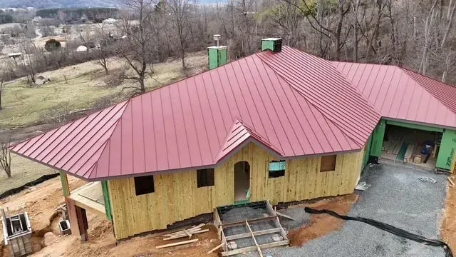 A house under construction with a red metal roof and wooden exterior. A driveway and surrounding landscape are visible.