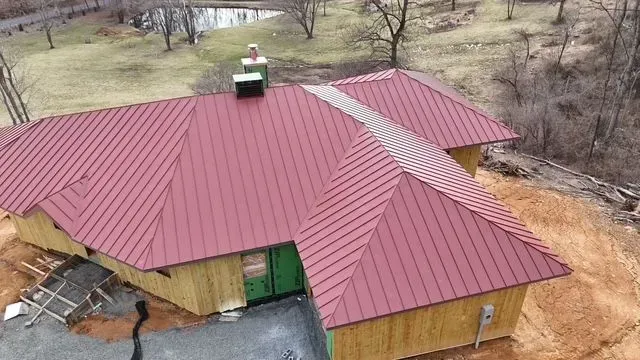 Red metal roof on a house under construction; surrounded by trees and a pond.