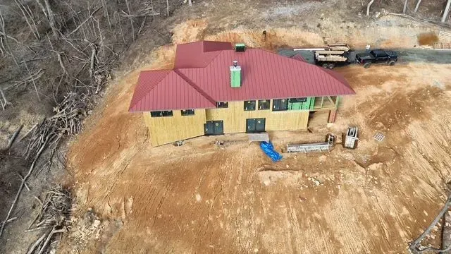 House under construction on a hillside, red metal roof, light brown walls, green chimney, and construction equipment.