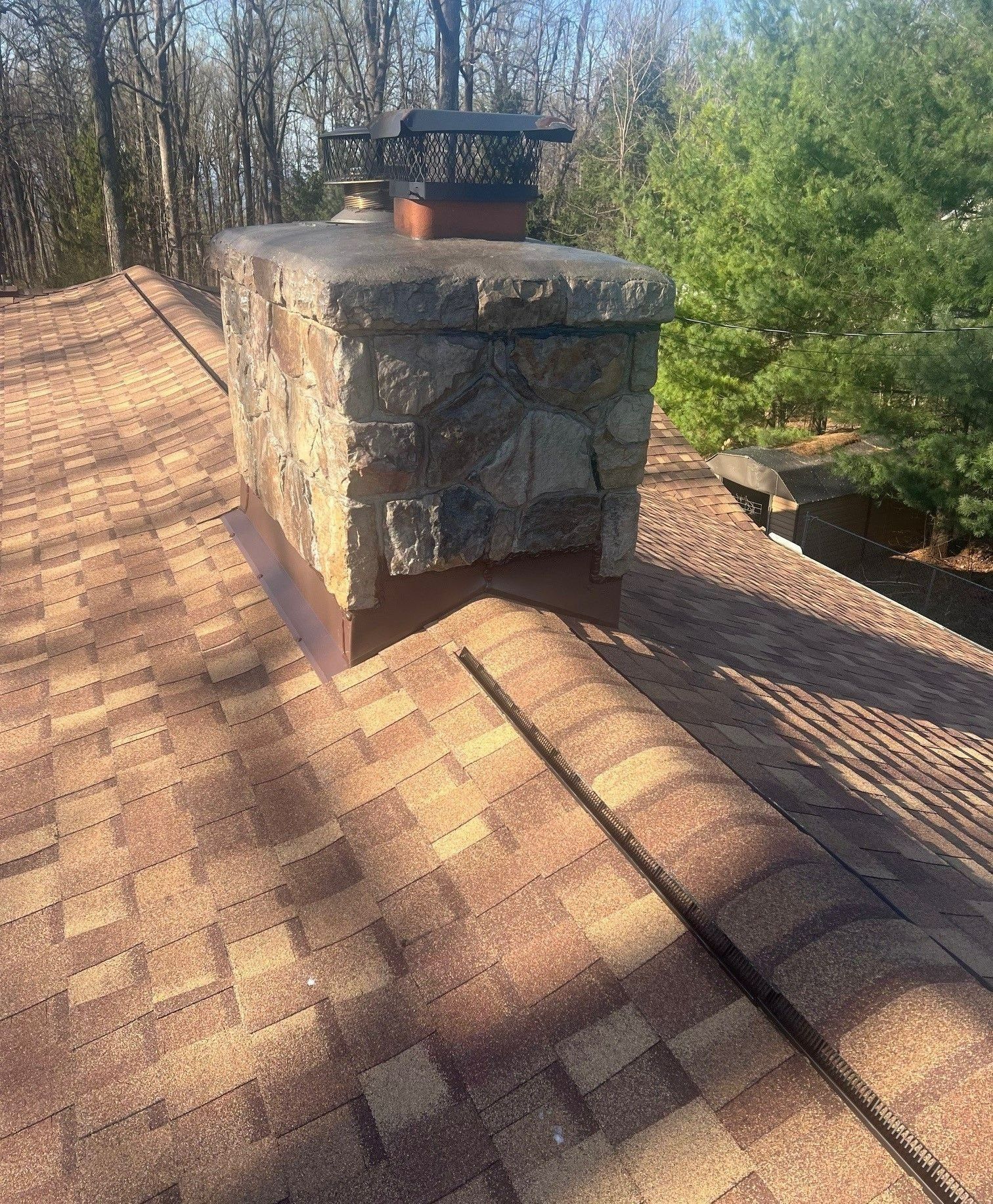 Stone chimney on a brown shingled roof with copper flashing, surrounded by trees.