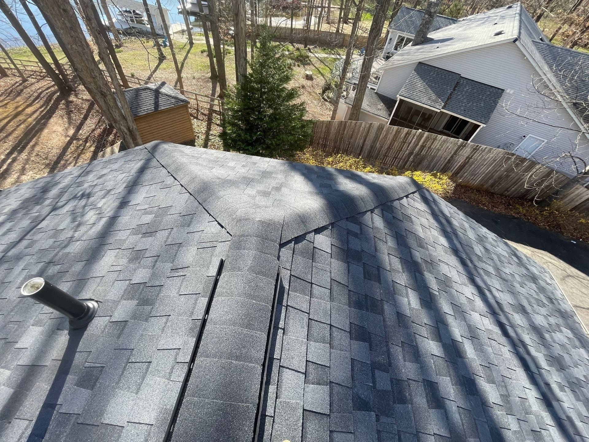 View from a roof; dark asphalt shingles, a vent pipe, trees, and houses in the background.