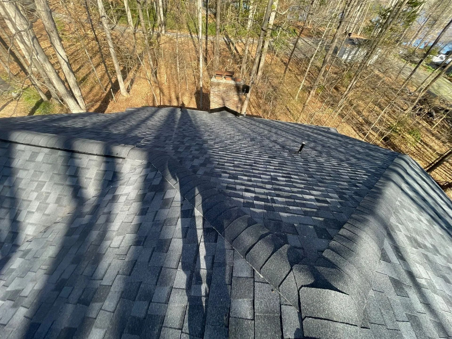 Overhead view of a dark shingled roof with chimney, trees in background. Sunny day.
