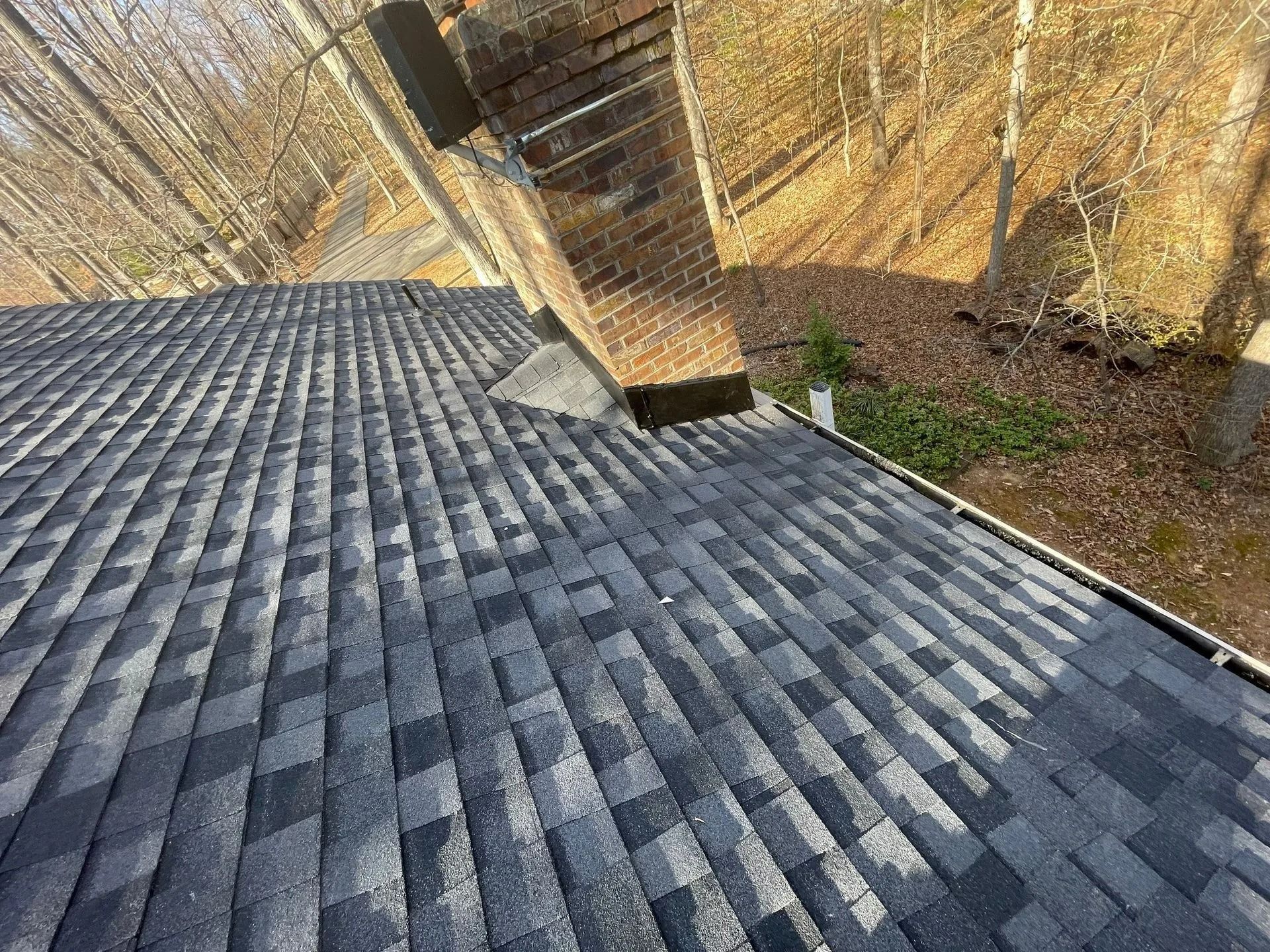 A newly shingled roof with a brick chimney against a backdrop of trees on a sunny day.