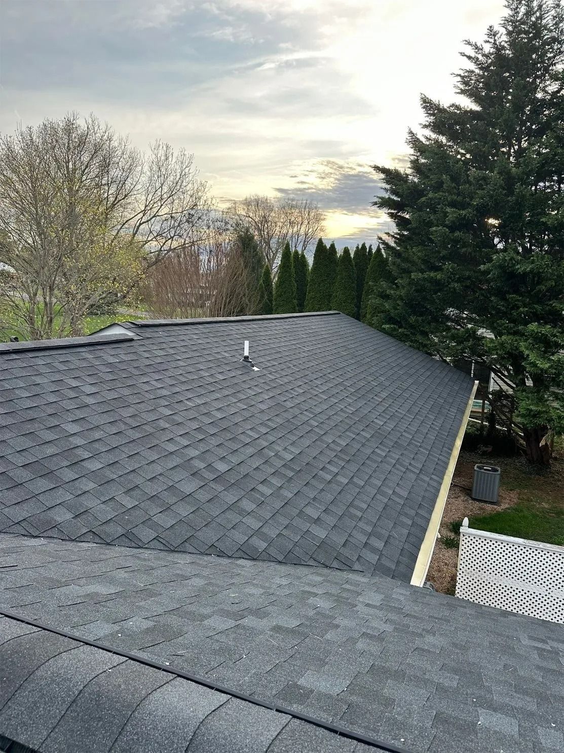 Dark gray shingle roof with a vent pipe against a cloudy sky, viewed from a higher angle.