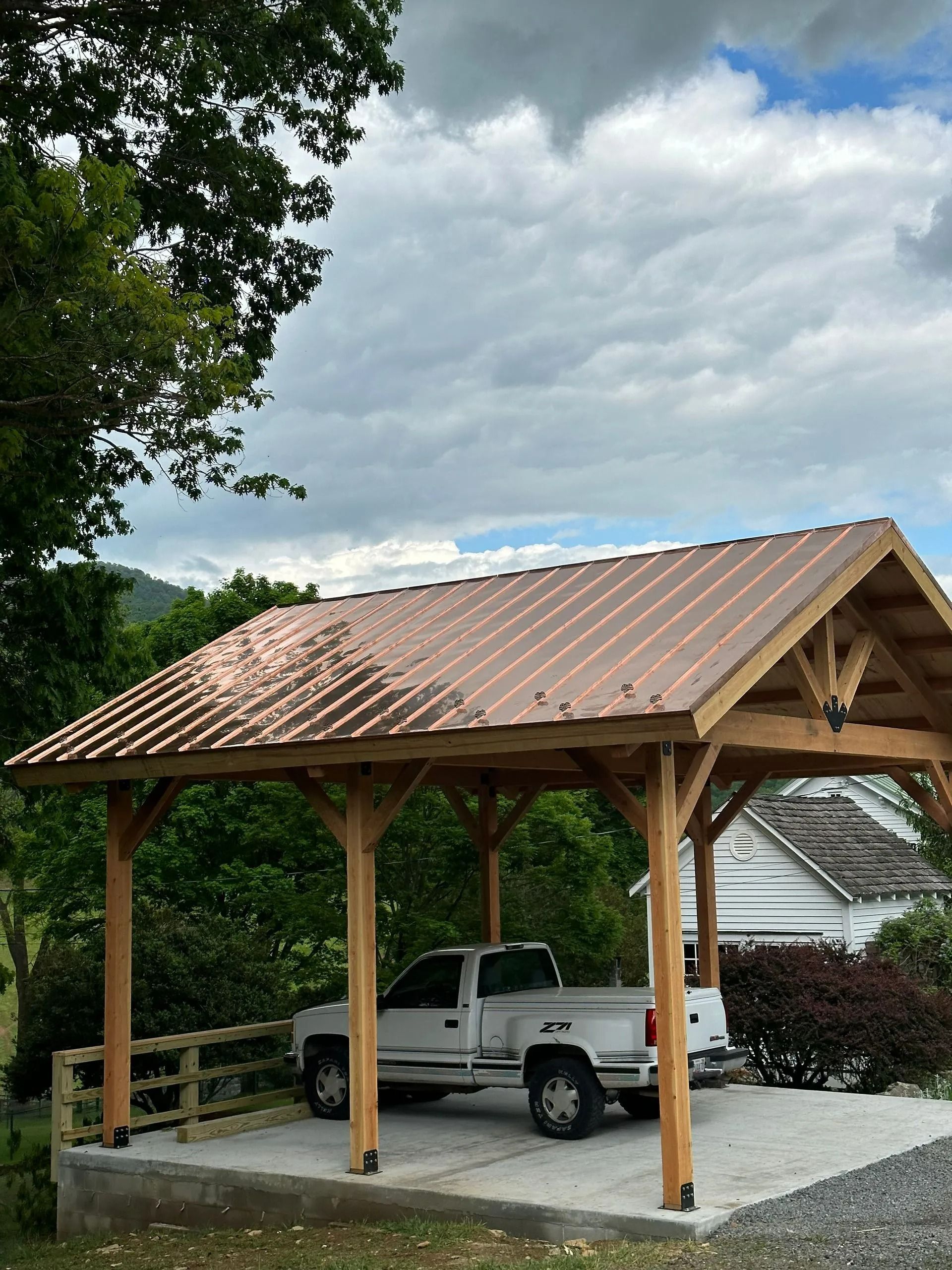 Carport with copper roof sheltering a white pickup truck. Wooden posts and frame, concrete pad, against a cloudy sky.