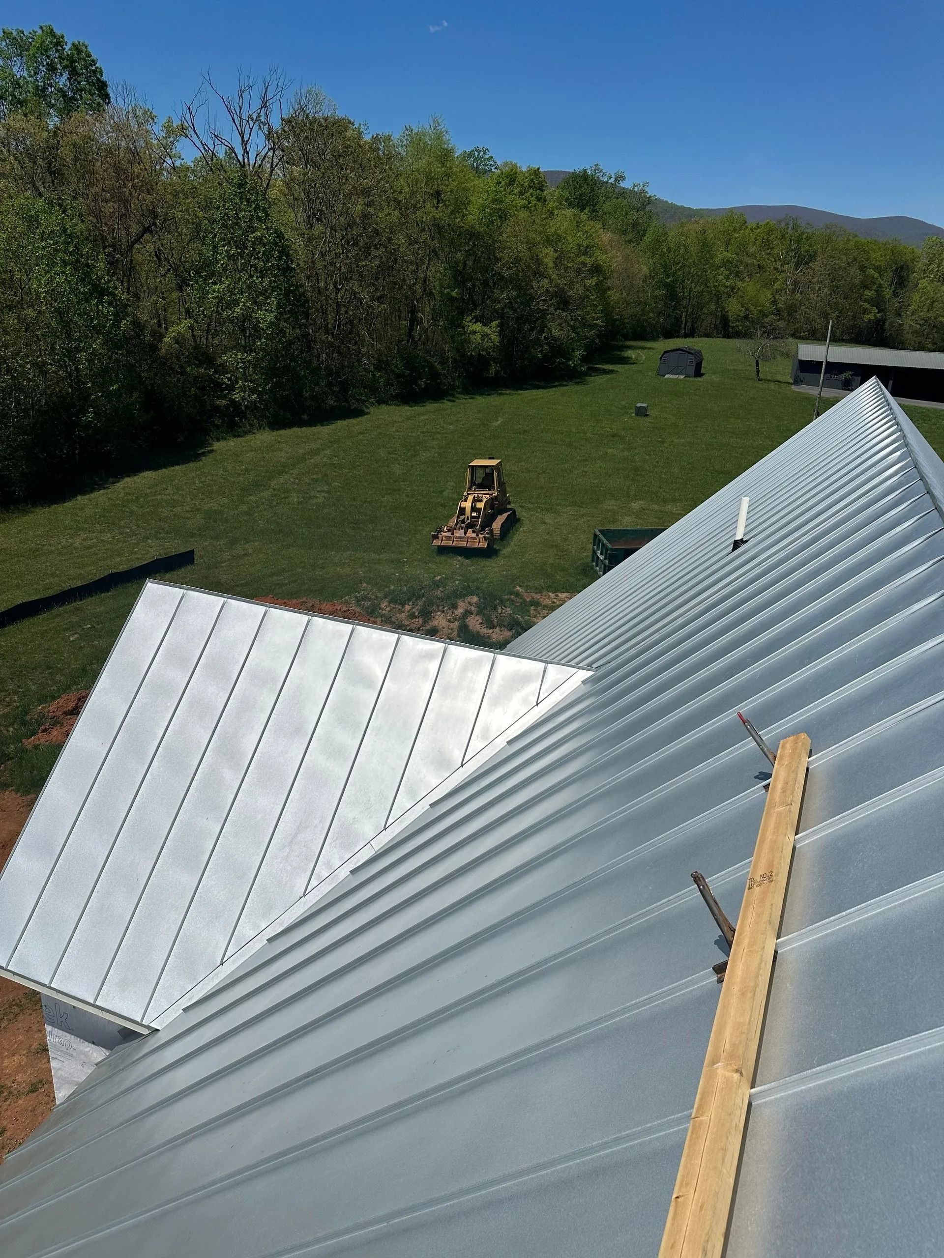 Metal roof installation on a sunny day; bulldozer on green grass, trees in background.