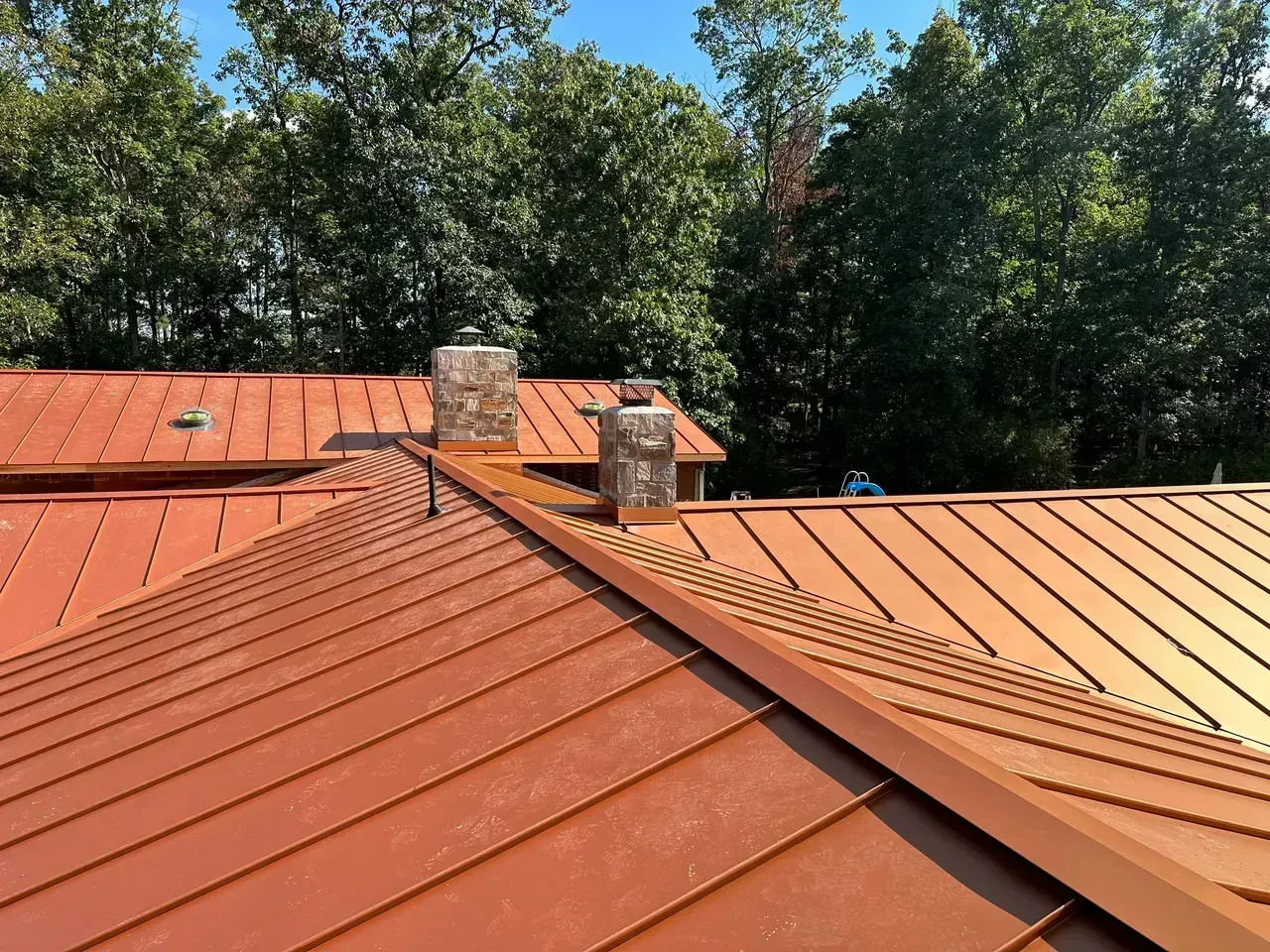 Copper-colored metal roof with two chimneys against a backdrop of green trees and a blue sky.