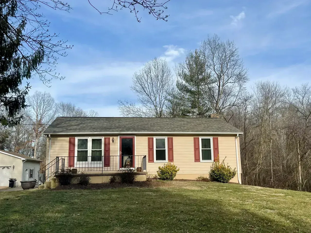 Tan ranch house with red shutters and a small porch, set on a grassy hill under a blue sky.