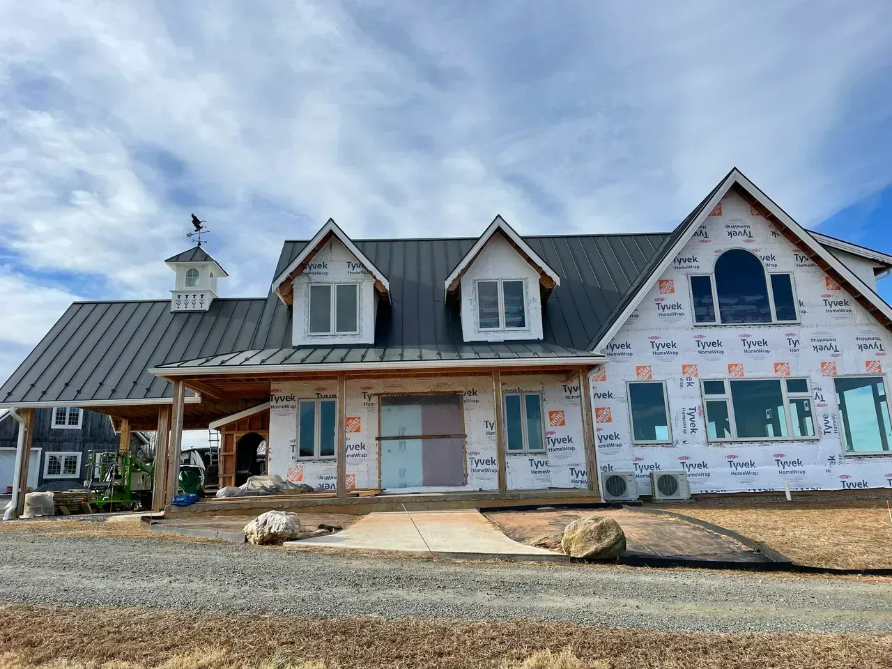 Barn-style house under construction with white siding, black roof, and unfinished windows, set against a blue sky.