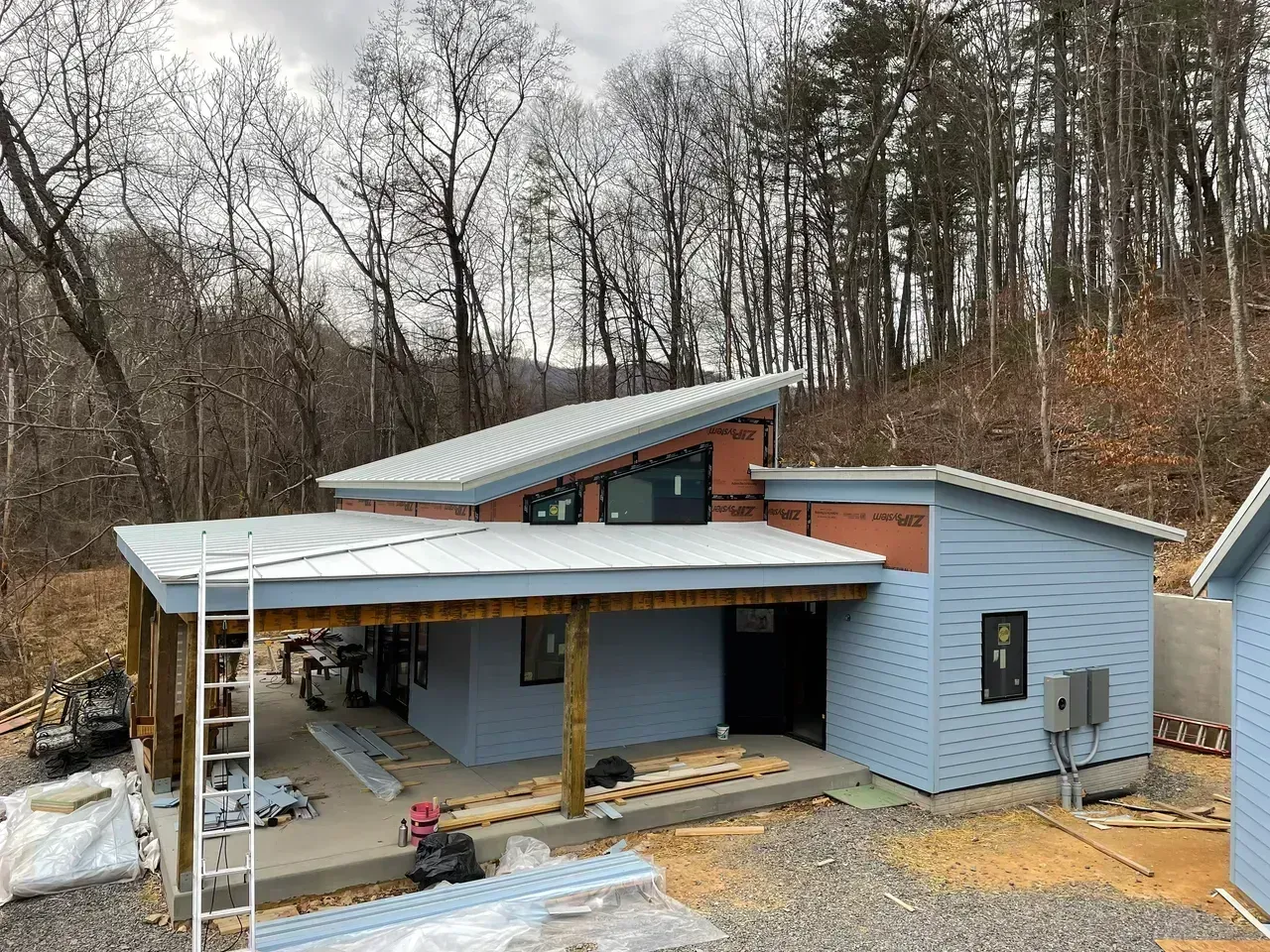 Blue-sided house with covered porch and angled roof under construction, set against a wooded hillside.