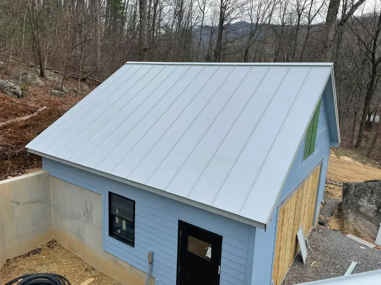 Small blue shed with a metal roof, black door and window, in a wooded setting.