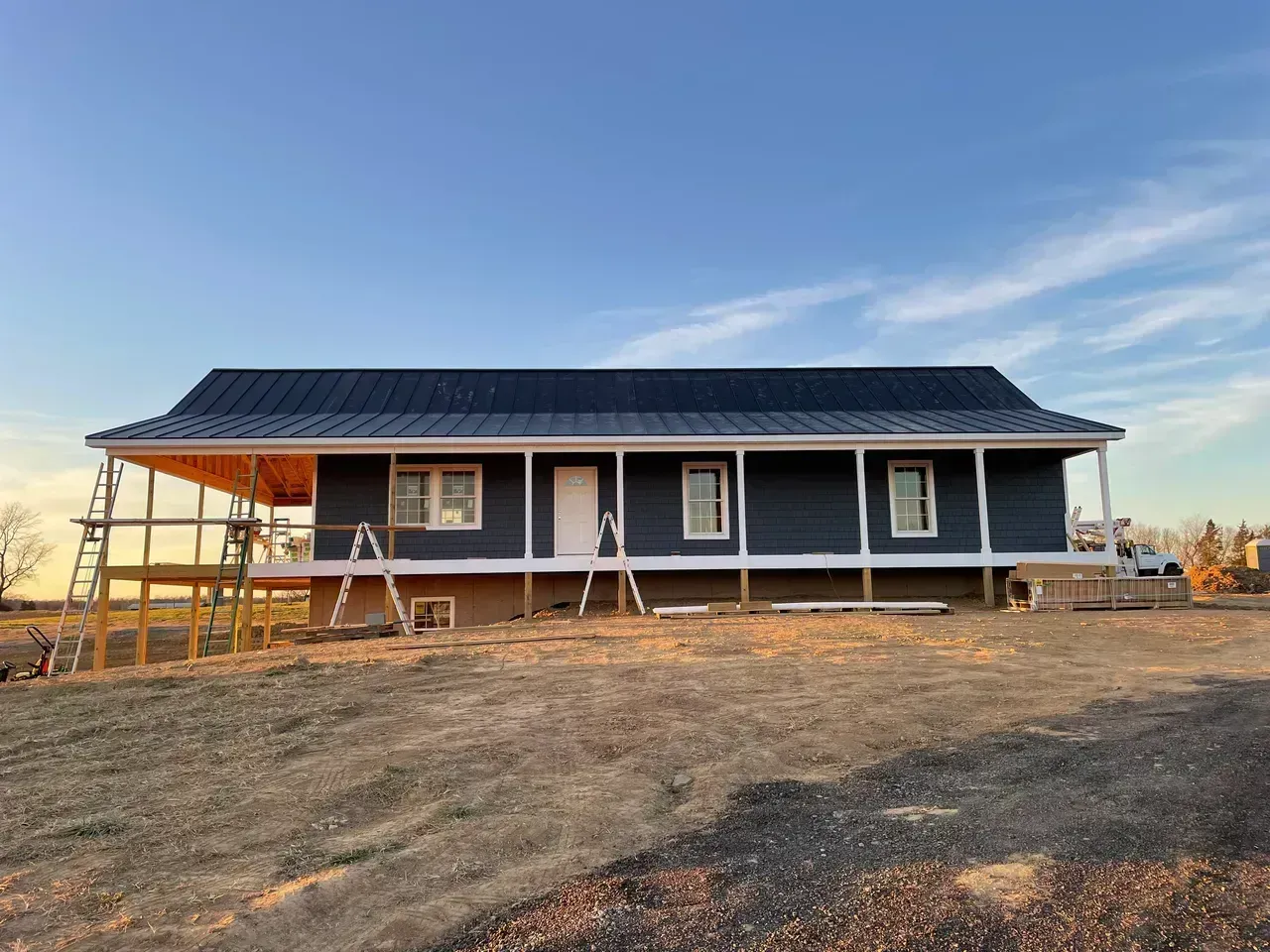 House under construction with dark blue siding, white trim, and a black roof. Ladders and wooden supports are visible.