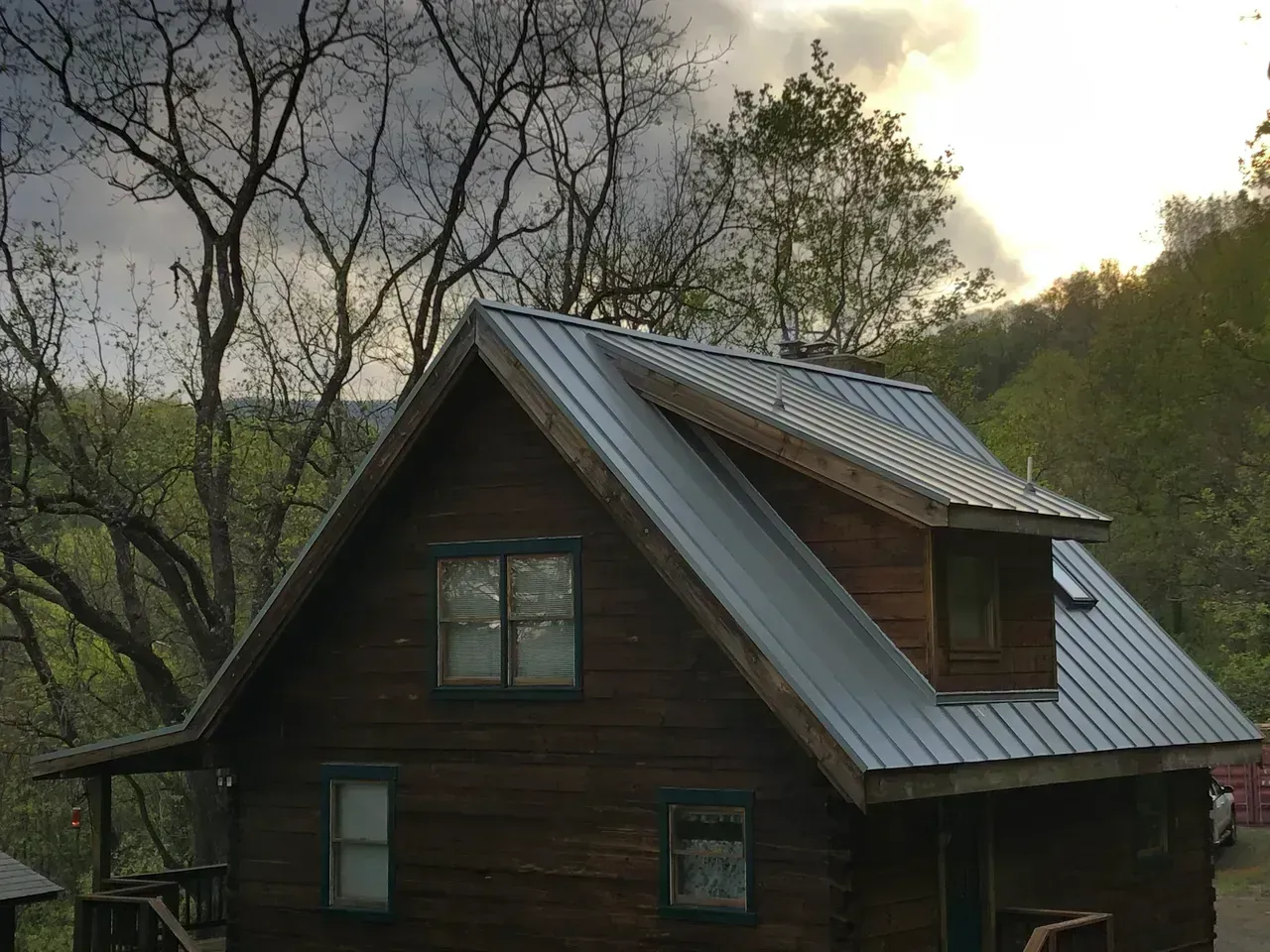 Log cabin with gray metal roof, windows, and porch, nestled in a wooded setting.