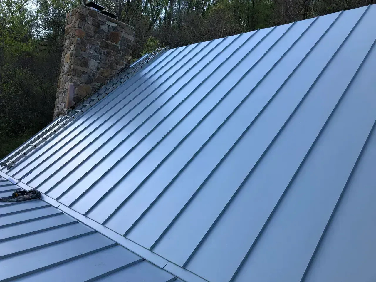 Metal roof with raised seams in a light gray color, chimney in the background.