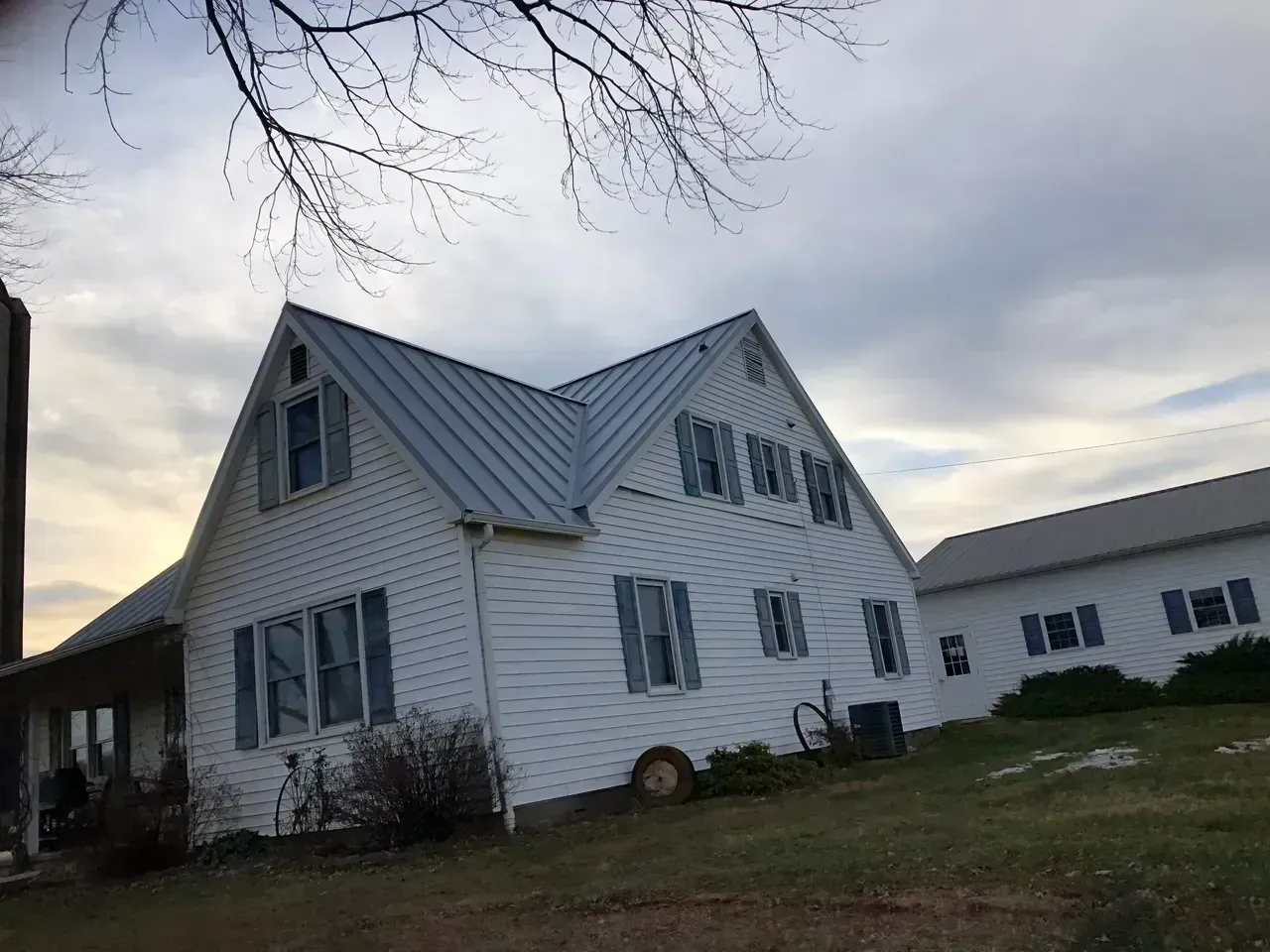 White farmhouse with blue shutters and metal roof on a cloudy day.