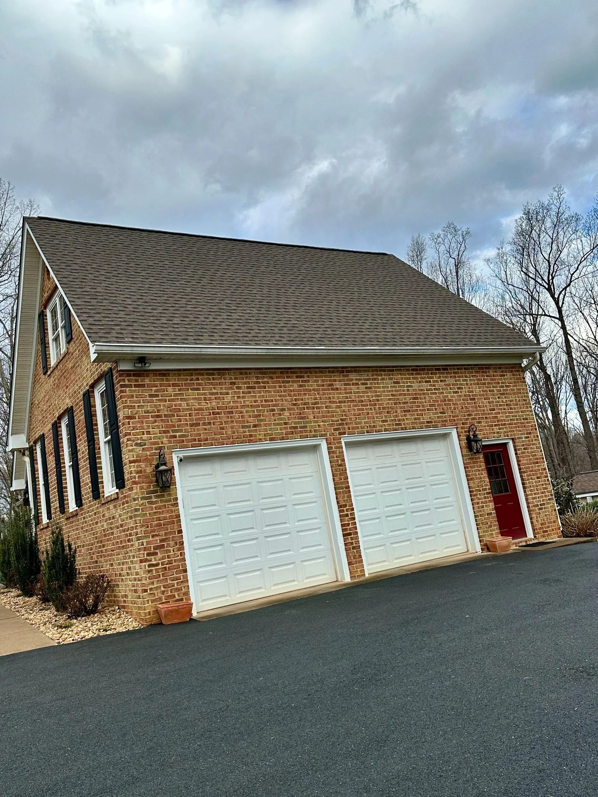Brick house with two white garage doors, a red door, and a dark roof under a cloudy sky.