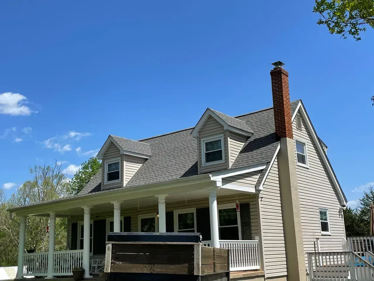 House with gray roof, white porch, tan siding, and brick chimney against a blue sky.
