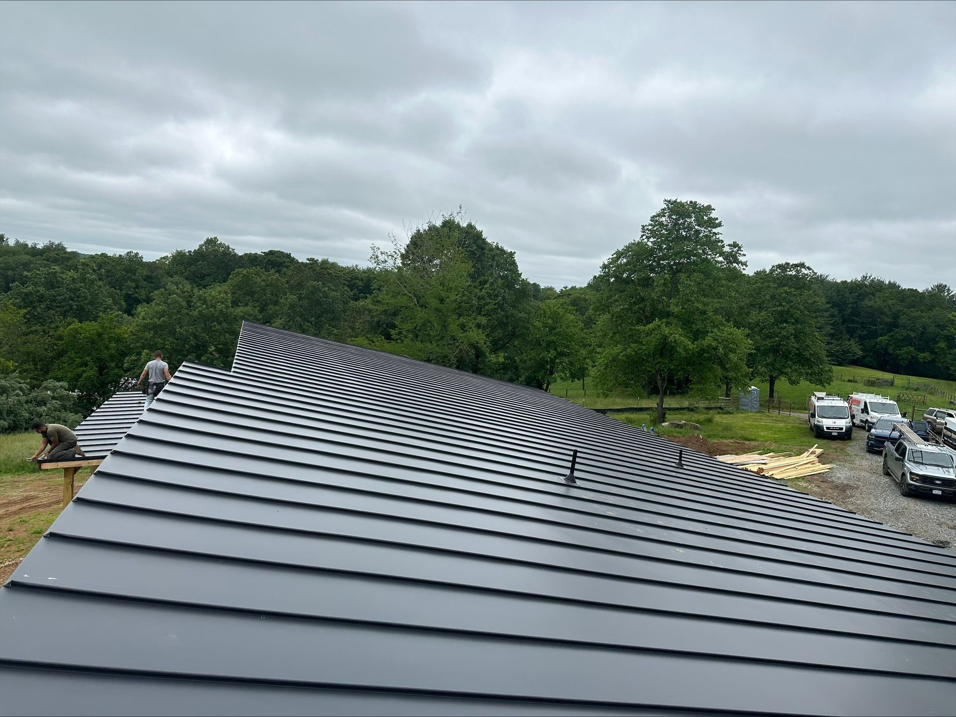 Dark metal roof with wavy lines, trees, and parked vehicles under a cloudy sky.