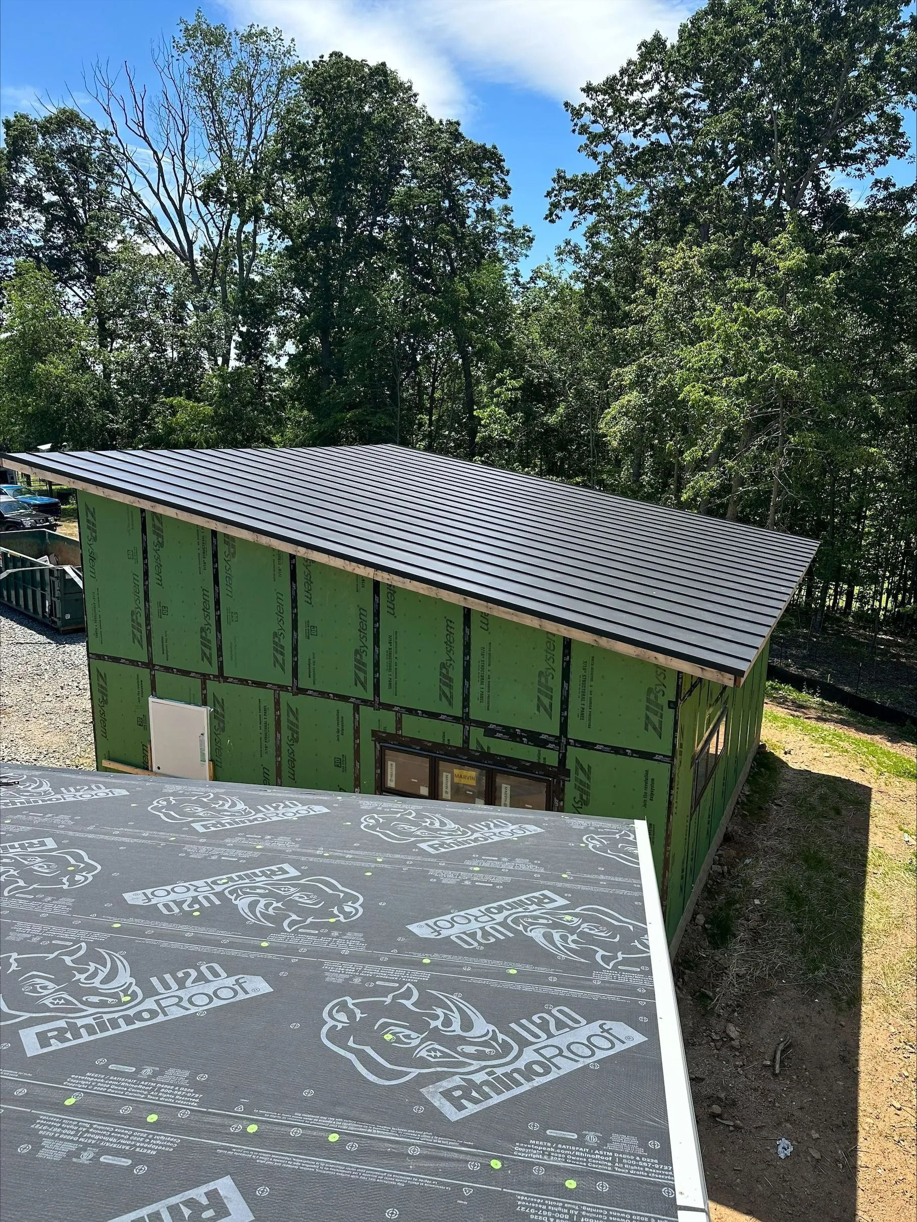 Green structure with angled roof, black tiles. Building site, trees.