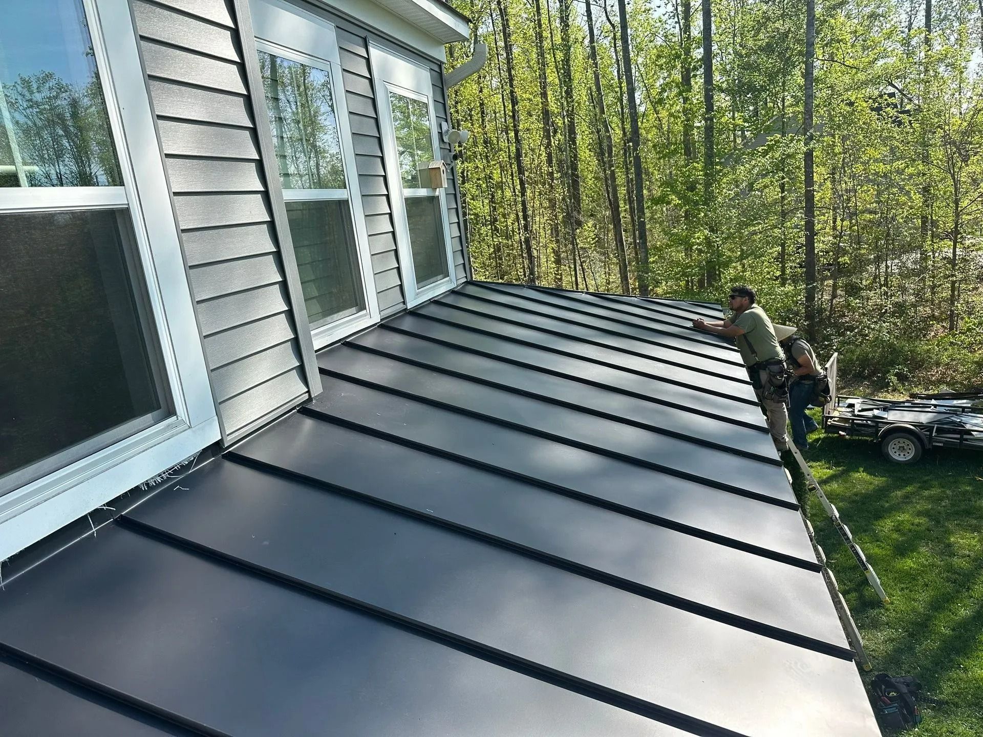 Two men installing a dark metal roof on a house with gray siding; green trees in the background.