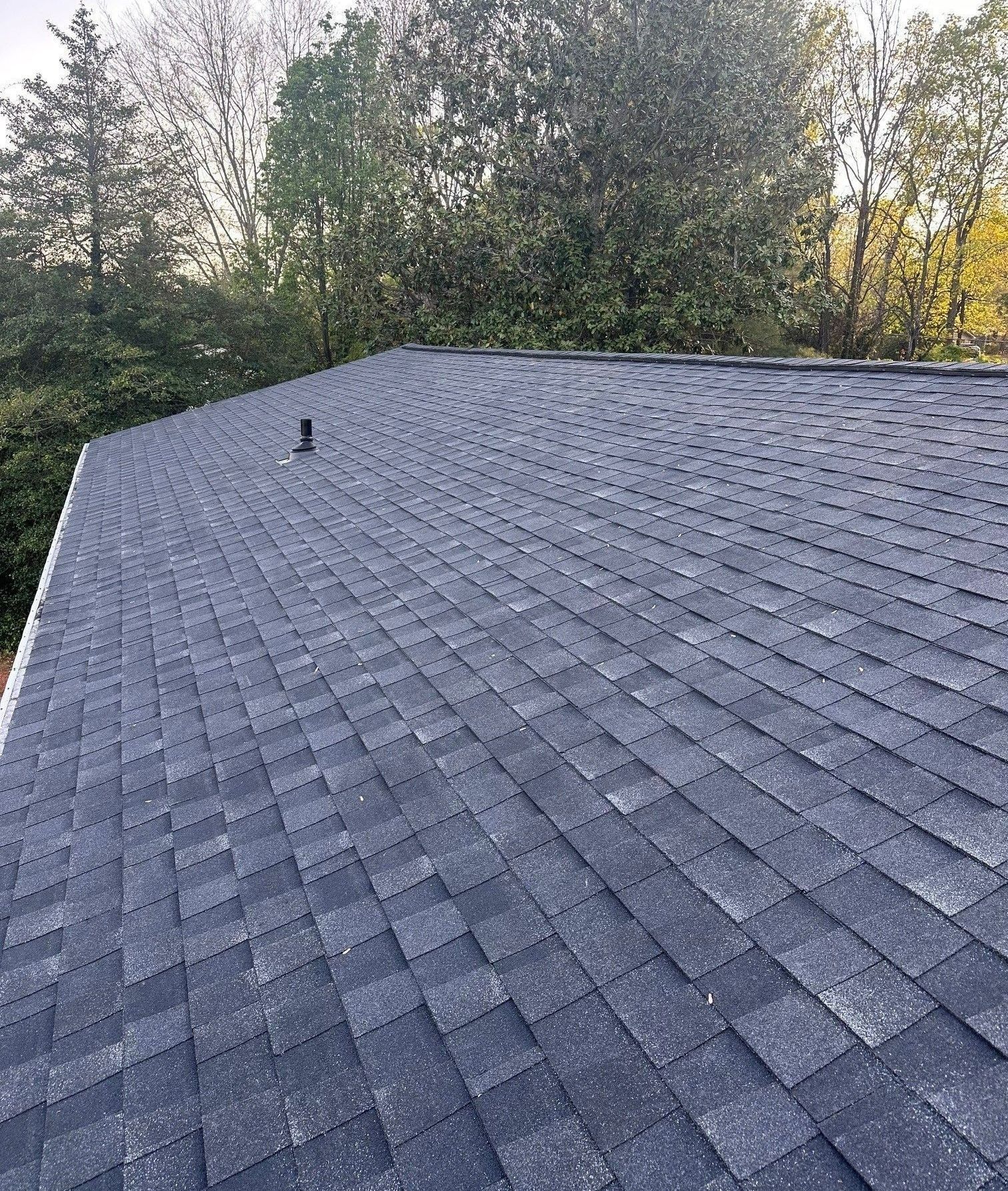 Dark blue asphalt shingle roof with a vent pipe, viewed against a backdrop of trees.