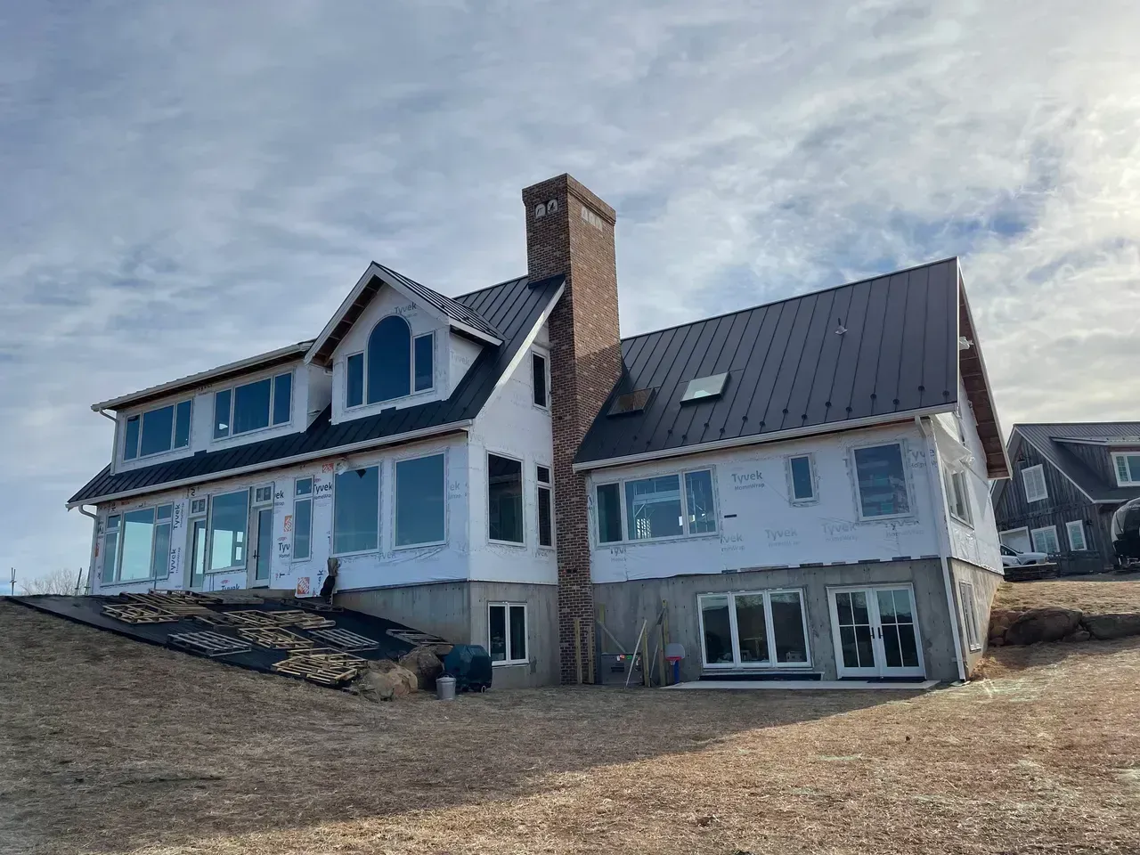 House under construction on a grassy hill. Black roof, brick chimney, large windows, and white siding are visible.