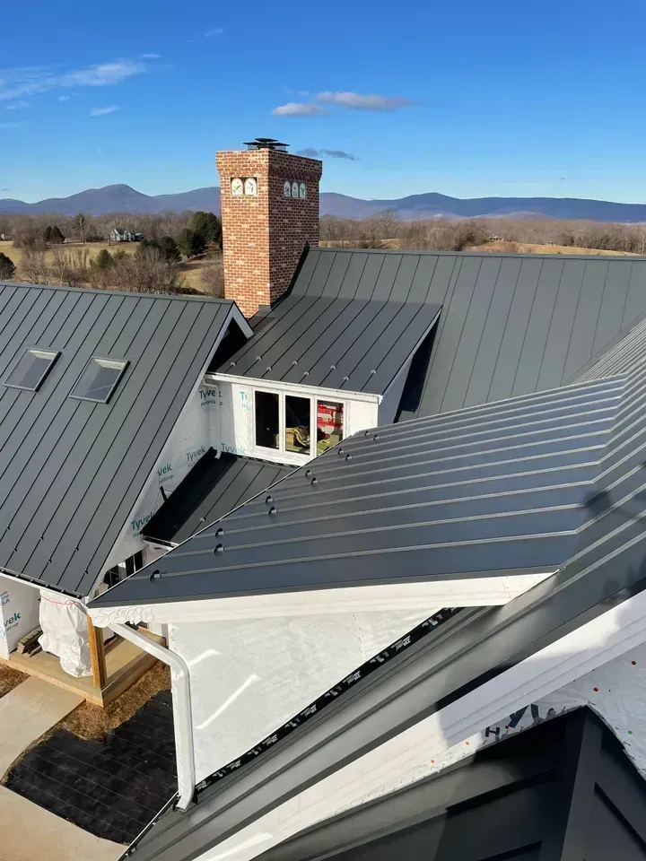Black metal roof on a white house with a brick chimney, against a mountain backdrop.