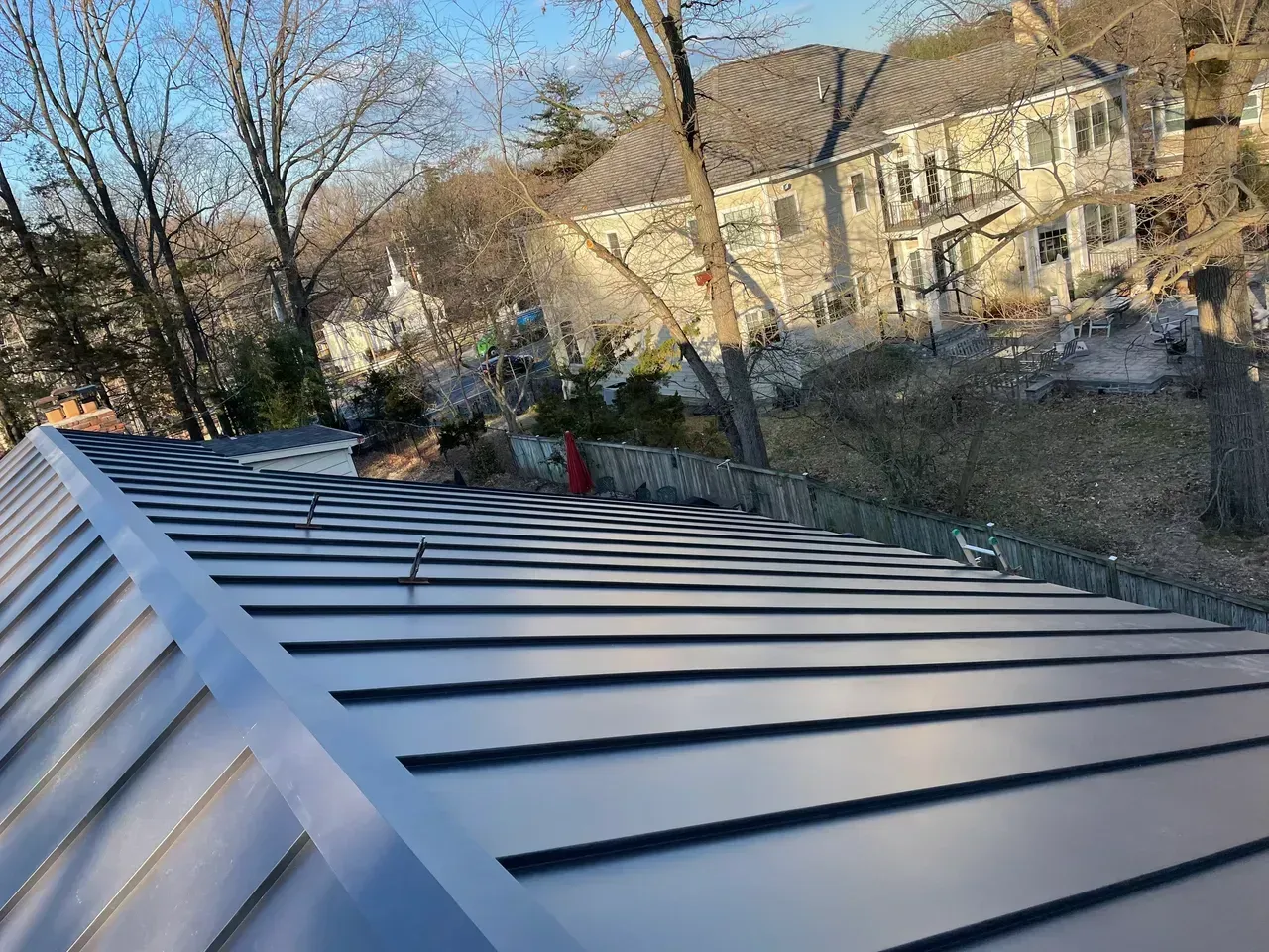 Gray metal roof with a large house in the background, trees and a fence in a sunny setting.