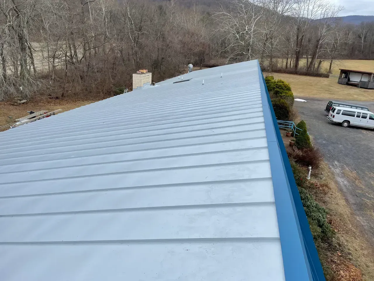 Metal roof with a blue edge on a building, surrounded by trees, a van, and other structures.