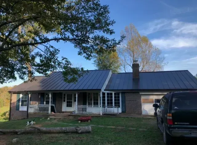 Ranch-style house with a dark blue metal roof, porch, and garage, on a grassy lot with a truck parked outside.