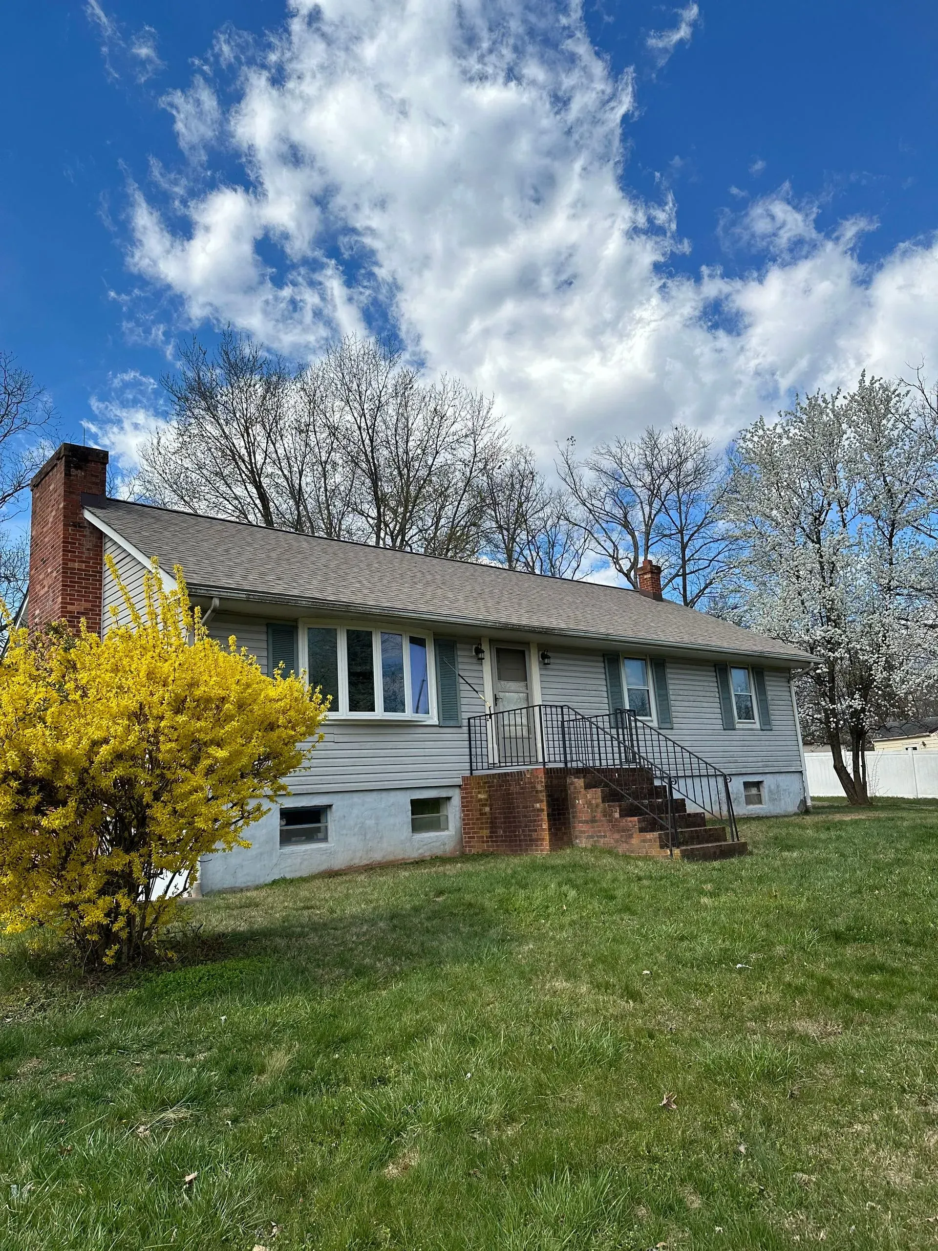 Ranch-style house with brick chimney, steps, and flowering trees on a sunny day with blue sky and clouds.