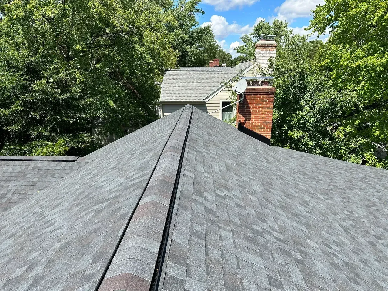 Gray asphalt shingle roof with a ridge, trees, and a brick chimney against a blue sky.