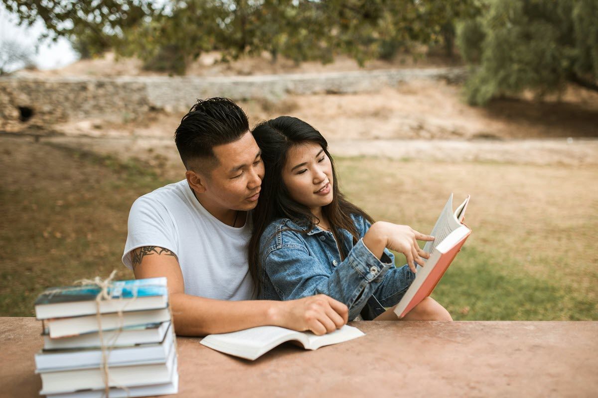 Couple reading books together outdoors at a table, pointing at pages. Stack of books nearby.